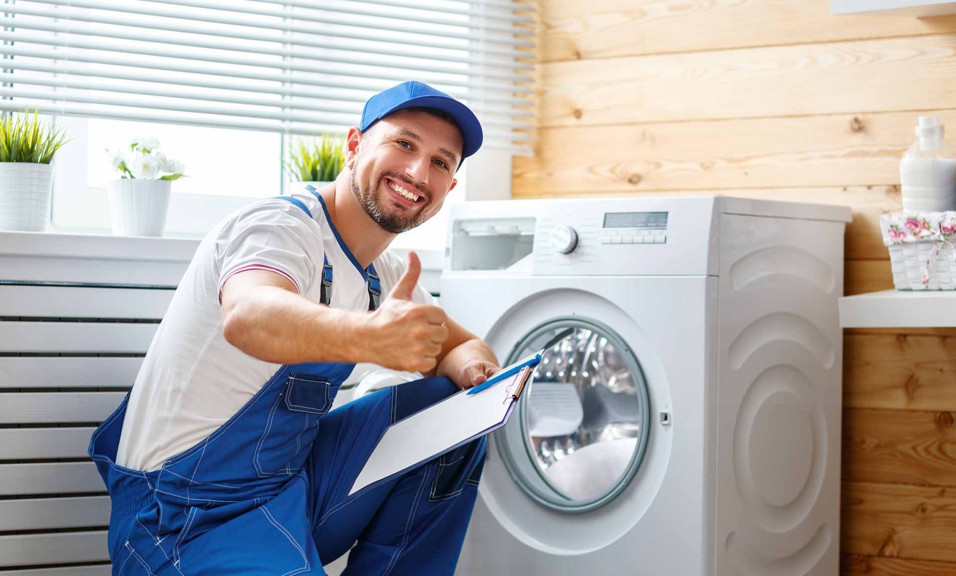 Appliance repair technician kneeling in front of a washer/dryer. He is smiling at the camera and giving a thumbs up gesture